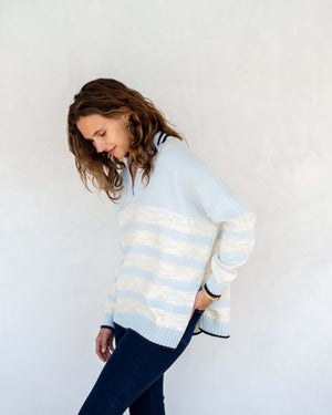 A person with shoulder-length wavy hair is standing against a light-colored wall, wearing the MERSEA - MARINA POLO SWEATER that exudes maritime spirit and dark pants. They are facing the camera with a relaxed expression as shadow patterns fall across the wall behind them.