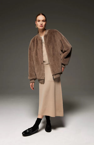 A person with tied-back hair is wearing the SEVENTY - MIDI KNIT SKIRT, an A-shaped beige midi-length skirt with an elasticated waist, paired with a matching beige top. The ensemble is complemented by an oversized brown fur jacket. Black socks and shiny black shoes complete the chic, minimalist look against a neutral grey background.