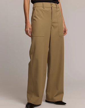 A person stands facing away, wearing HINSON WU's KELLY TRAVELER'S TWILL PANT—high-waisted, wide-leg tan trousers with large back pockets—paired with a black top against a light neutral background.