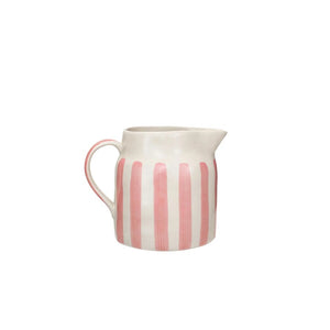 Two Creative Coop 1 3/4 Quart Pitchers with vertical stripes, one featuring blue and orange hues and the other in pink, rest elegantly on a stack of books atop a light-colored surface. The neutral background ensures that these vibrant hand-painted stoneware pitchers take center stage.