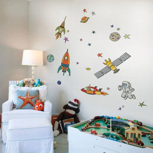 A whimsical space scene featuring colorful rockets, an astronaut, and various planets, stars, and a satellite. The SIMPLE SHAPES - OUTER SPACE REMOVEABLE WALL DECALS by SIMPLE SHAPES showcase fun designs and colors on the rockets, while the astronaut appears to be floating in a spacesuit near Earth and other celestial objects.