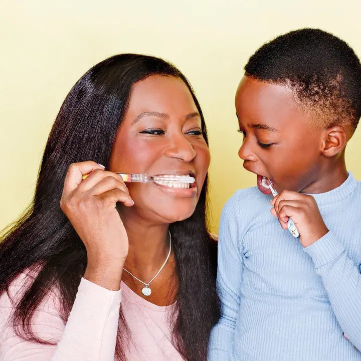 A woman with long hair and a young boy are smiling while brushing their teeth together using HAMICO - KIDS AIRPLANE TOOTHBRUSH. The woman, demonstrating proper brushing techniques with the dentist-designed toothbrush from HAMICO/INNOVATIVE TOOTHBRUSH, holds it to her mouth. The boy enthusiastically imitates her actions, all set against a soft yellow background.