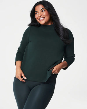 A person with long, dark hair smiles and poses with one hand in their pocket. They are wearing the SPANX - AIRESSENTIALS MOCK NECK TOP made from super-soft fabric, paired with matching dark green leggings against a light background.