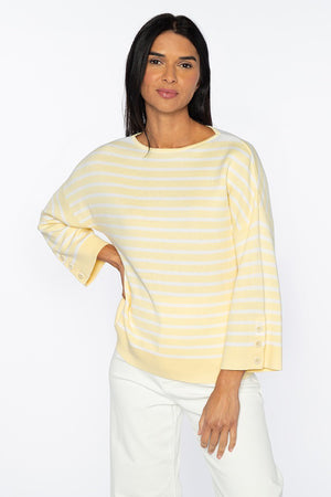 A woman stands against a white background, wearing the KINROSS CASHMERE - STRIPE BUTTON SLEEVE PULLOVER from KINROSS. The light yellow and white striped, loose-fitting sweater is made of soft cotton and features three-quarter sleeves with button details. She pairs it with white pants. She has long dark hair and is smiling softly at the camera.