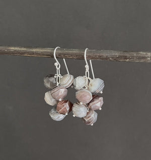 A pair of ANN LIGHTFOOT - Faceted 9mm Botswana Agate Droplet Earrings features clusters of gray and brown beads, creating a textured look. They hang from a dark rod against a plain white background.