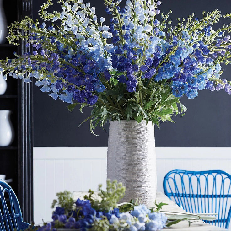 Introducing the DELPHINIUM STEM by NAPA HOME AND GARDEN: A single stem of vibrant purple-blue delphinium flowers with green leaves, arranged against a plain white background. The realistic blossoms are clustered along the upper part of the stem, while the decorative green leaves add a touch of natural elegance to elevate your home decor.