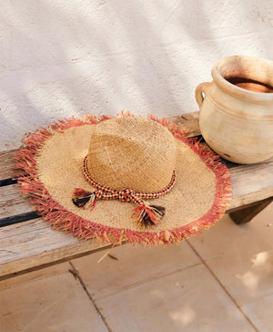 Introducing the TALLULAH RAFIA SUMMER HAT by MOMONI: This exquisite natural raffia hat boasts a wide, fringed brim with a distinctive frayed blue edge. Adorned with an elegant blue and white chenille and lurex hatband tied around the base of the crown and accented by a delicate tassel, this hat is showcased against a plain white background.