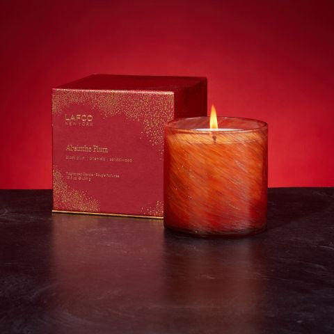 A glowing, orange-colored candle made from soy wax is placed next to a red box labeled "LAFCO - ABSINTHE PLUM HOLIDAY CANDLE 15.5OZ." The scent of black plum and sandalwood permeates the air. The candle's warm, inviting glow is beautifully enhanced by the rich red background.