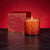 A glowing, orange-colored candle made from soy wax is placed next to a red box labeled "LAFCO - ABSINTHE PLUM HOLIDAY CANDLE 15.5OZ." The scent of black plum and sandalwood permeates the air. The candle's warm, inviting glow is beautifully enhanced by the rich red background.