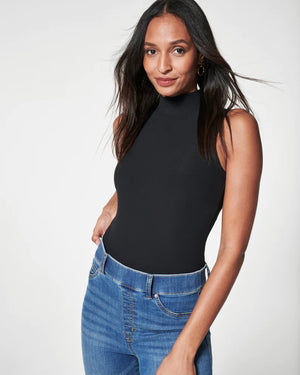 A woman stands confidently against a plain white background, wearing a fitted black leather jacket, a black top, and SPANX - FLARE JEANS IN VINTAGE INDIGO. She has long dark hair and is looking directly at the camera with a slight smile. Her left hand is in her pocket.