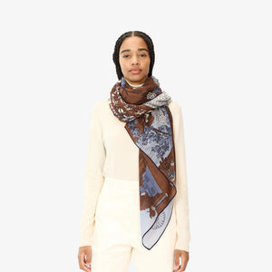 A person stands wearing the INOUI EDITIONS - QUATRE SAISONS SCARF, a long and lightweight summer accessory featuring detailed, intricate designs in brown, blue, and white tones, draped over a cream-colored top. The unique and colorful scarf from INOUI hangs elegantly around the neck and over one shoulder against a plain white background.