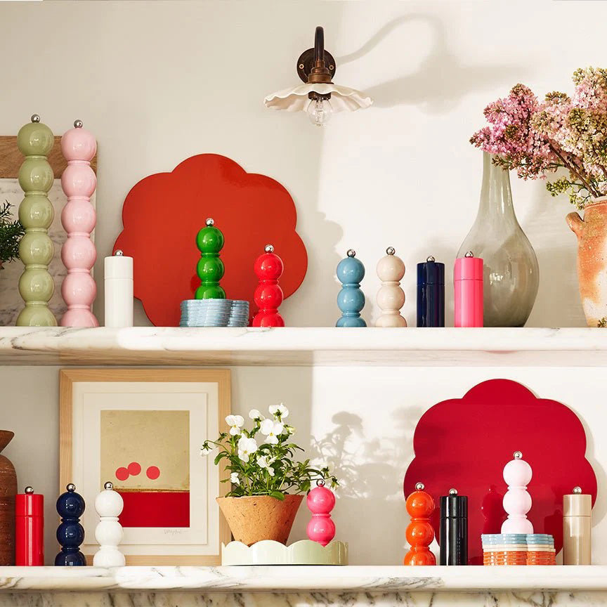 A kitchen shelf displaying colorful salt and pepper grinders, including an Addison Ross London Mini Bob Salt or Pepper Mill (14cm) with a ceramic mechanism, a potted plant with white flowers, and various decorative items. There are two framed pieces of art, vases with flowers, red and orange placemats, and a wall-mounted light.