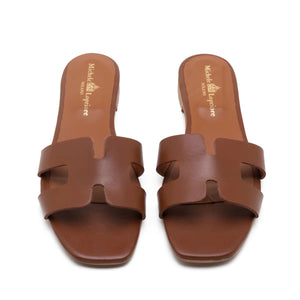 The image depicts the MICHELE LOPRIORE - H SLIDE, a single brown leather slide sandal with an open-toe design. The upper features two wide straps with geometric cutouts. The thin sole appears to be made of light brown wood-like material. Made in Italy by MICHELE LOPRIORE, the sandal is displayed against a plain white background.