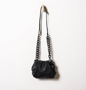 A black, textured handbag with a rectangular base and a gathered top is displayed on a white pedestal. The handbag, designed by DANIELA LEHAVI and named the TOKYO BALI BAG, features an alligator-like pattern and appears to be made of leather or a similar material. The background is plain and neutral-colored.