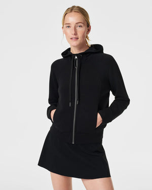 A person with blonde hair is wearing the SPANX AirEssentials Full Zip Hoodie made from ultra-soft fabric, with their hands in the pockets and a black skirt. They are standing against a plain, light-colored background and smiling softly.