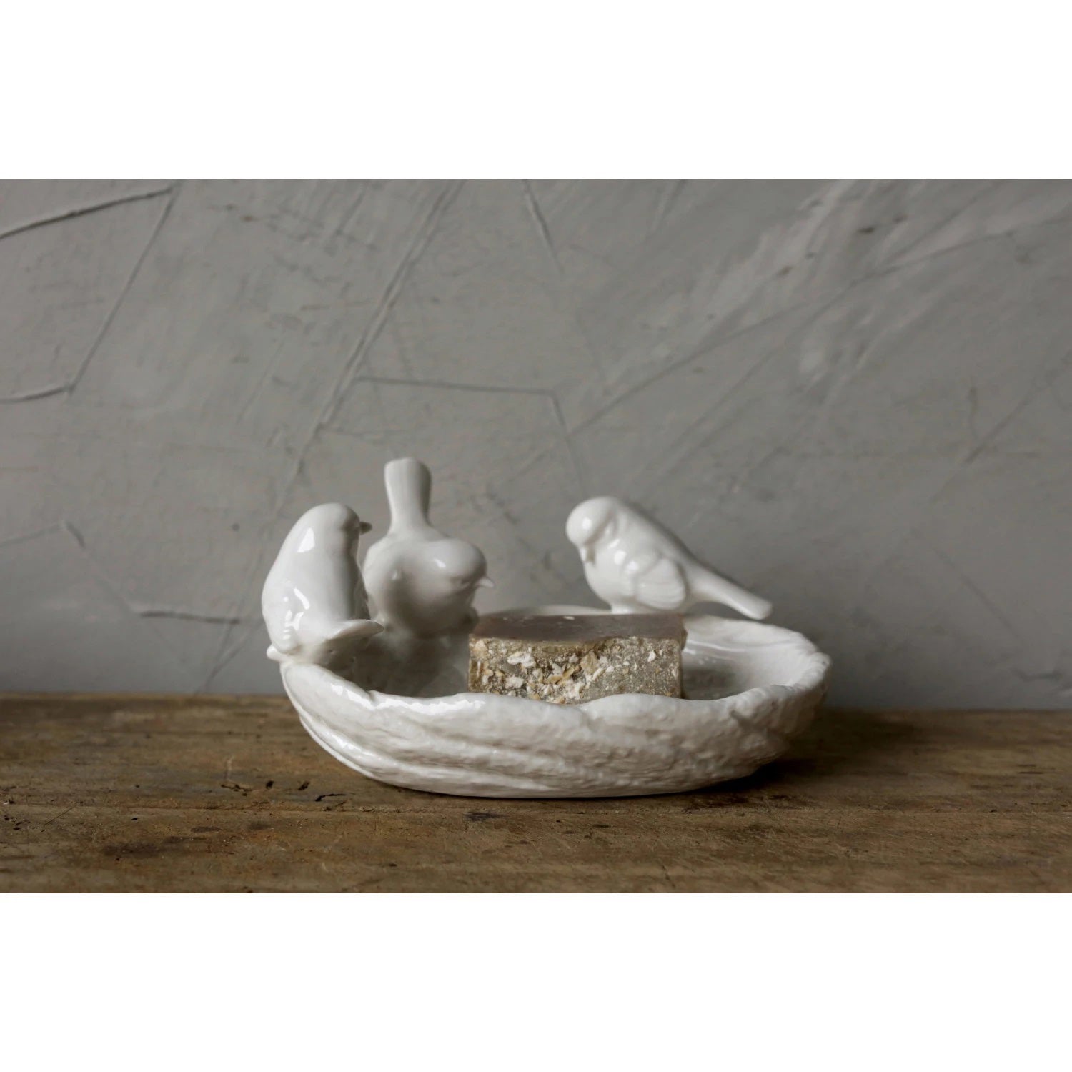 The LEAF DISH WITH BIRDS by CREATIVE COOP is a white ceramic trinket holder designed to resemble a bird bath, featuring three birds perched on the edge. The surface has intricate detailing that mimics the texture of a real bird bath.