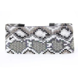 Introducing the JULIE ROFMAN - SNAKE CUFF by JULIE ROFMAN JEWELRY: This bold and intricate bracelet is adorned with black, gray, and white glass delica beads in a striking patterned design. Handwoven with elements reminiscent of snakeskin, it features a black buckle closure and leather-like ends, expertly combining geometric and textured elements for a truly unique style.