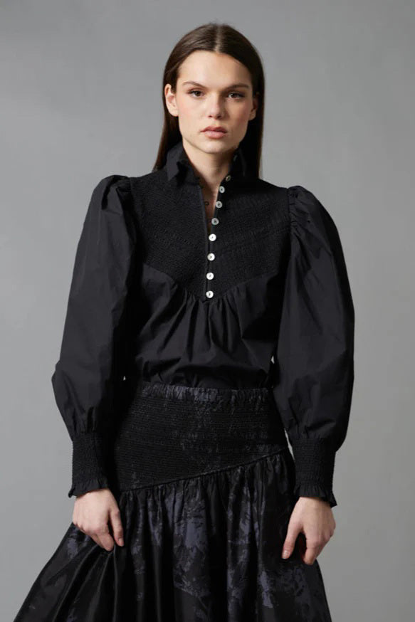 A woman with long brown hair faces away, wearing the HOPE FOR FLOWERS Smocked Shirt in Black with puffed sleeves and paired with a dark floral skirt, standing before a gray backdrop.