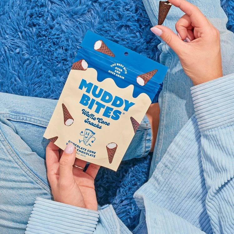 A person dressed in a light blue outfit sits on a plush, royal blue rug holding a bag of Muddy Bites Cookies & Cream Waffle Cones in one hand and a small white chocolate waffle cone snack in the other. The beige and blue bag features an illustration of chocolate-dipped waffle cones.