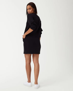 A woman is wearing the SPANX AirEssentials Crew Neck Dress, a black, long-sleeve, knee-length dress from the SPANX brand, with her hands in the functional pockets. She is looking to the side and has long, straight hair. Paired with white sneakers, she stands against a plain white background in this light-as-air fabric attire.