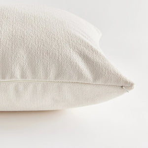 A close-up side view of the COOPER SQUARE INDOOR-OUTDOOR PILLOW 20" by NAPA HOME AND GARDEN against a plain, light background. The image showcases the pillow's white, textured Jacquard fabric and stitching detail, with the zipper partially visible on the edge.