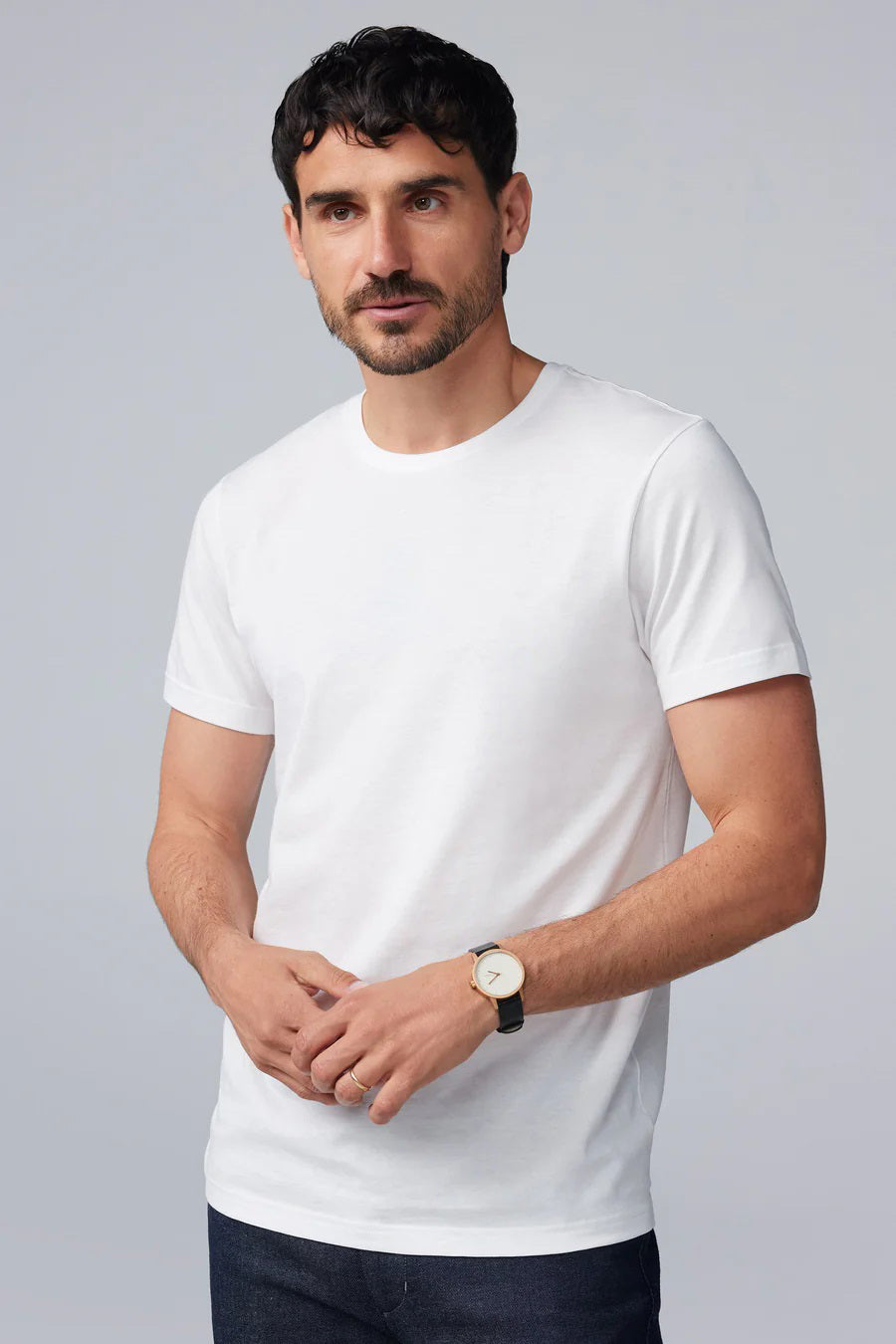 A man with short dark hair and a beard wears the MACK WELDON SILVER CREW NECK T-SHIRT IN WHITE and dark pants, standing in profile with his head slightly bowed and one hand in his pocket against a light gray background.