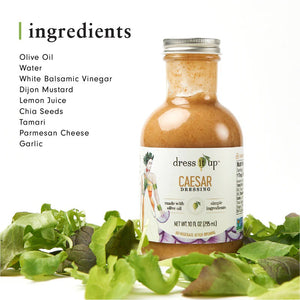 A 10 fl oz (295 ml) bottle of Dress It Up Dressing - Caesar dressing. The label showcases an illustration of a person in a green dress and emphasizes the product's low-calorie formulation made with olive oil and chia seeds, highlighting its simple, wholesome ingredients. The bottle is topped with a metal screw cap.