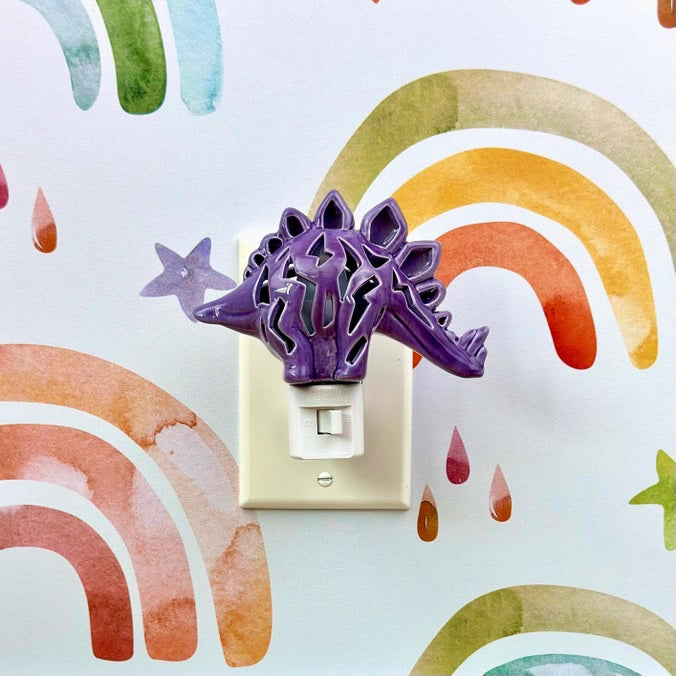 The DINOSAUR NIGHTLIGHT by LILY&#39;S LIGHTS is a purple ceramic stegosaurus-shaped nightlight that emits a warm glow through its cutout patterns. This handmade creation boasts quality craftsmanship, featuring a spiked back and standing on a white plastic plug base, suitable for standard electrical outlets. It&#39;s perfect for unique gifts.