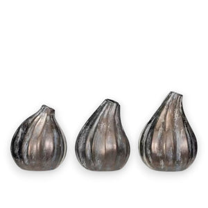 Three STONEWARE FIG SHAPED VASES by CREATIVE COOP in rustic, earthy tones are arranged on a neutral surface. Each vase features a reactive glaze finish, with one displaying a sprig of dried leaves. The scene is set against a subtly textured wall in the background.