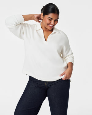 A person with dark hair styled in a neat updo is wearing the SPANX - AirEssentials Split Neck Polo Top in black, paired with matching black pants. They rest one hand on the back of their neck and gaze to the side against a plain white background, exuding an air of luxurious comfort.