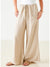 A person wearing a white sleeveless top and MERSEA's Provence Palazzo Pant by MER SEA, featuring a bohemian style with drawstrings and large front pockets. They pair it with white sandals while standing against a white brick wall.