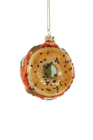 A CODY FOSTER BAGEL WITH LOX ORNAMENT, crafted from glass and detailed with sesame seeds and a green string, rests on green and white checkered paper resembling a sandwich with clear fillings. The paper showcases text and patterns in green, subtly accentuating the charm of this distinctive ornament.