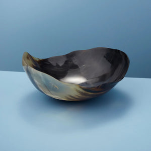 The HORN WAVY ROUND BOWL by BE HOME, INC, ethically crafted from water buffalo horn, showcases a polished, reflective surface. Its irregular, organic shape rests on a flat blue background. The natural blend of black and tan colors highlights the bowl's unique texture and sheen.