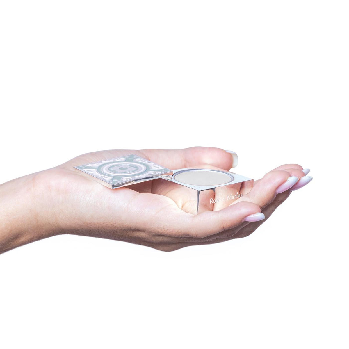 A manicured hand holds an open ROSE ET MARIUS - SUNLIT ROSE WATER SOLID PERFUME compact with a round pan, evoking subtle vanilla notes, against a white background.