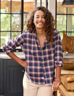 A person with wavy hair, dressed in a relaxed fit blue and red plaid Eileen shirt from Frank & Eileen, stands in a kitchen and glances over their shoulder. The kitchen features large windows, dark cabinets, and a wooden countertop with a bowl of fruit in the background.