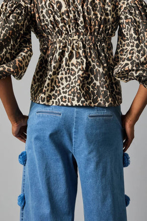 A person stands confidently against a gray background, wearing a geometric-patterned top with puffed sleeves and HOPE FOR FLOWERS' POM POM TROUSER made from organic cotton denim, featuring blue pom-pom accents along the sides. They complete the look with red sandals.