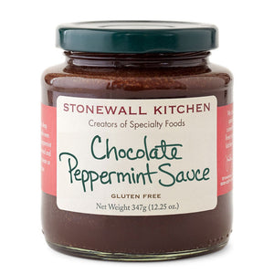 A jar of Stonewall Kitchen's Chocolate Peppermint Sauce, with a spoon inside, rests on a white tray. It's encircled by festive cookies and a cup brimming with peppermint candies, set against lush greenery and holiday decor—ideal for any dessert spread.