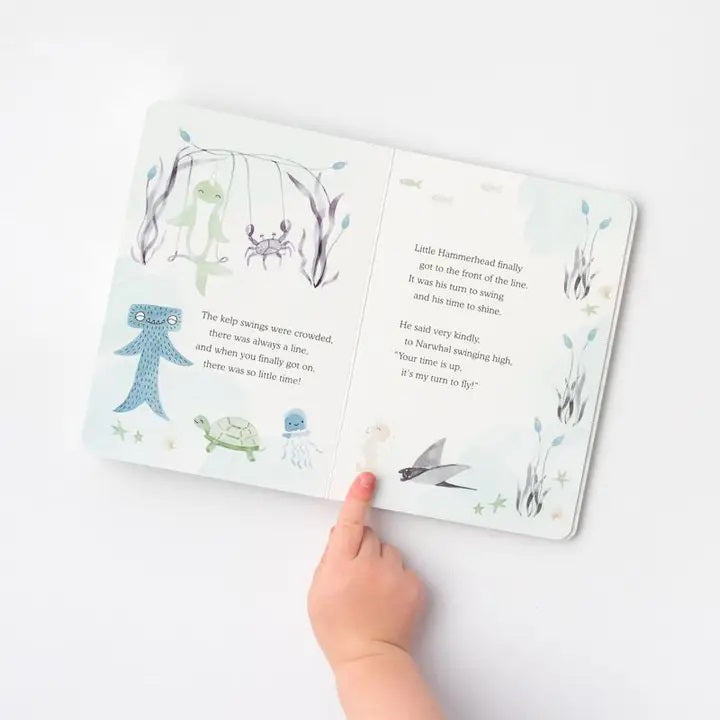A child&#39;s hand points at a page of the SLUMBERKINS - RECESS CHALLENGE BOOK by SLUMBERKINS. The page features illustrations of underwater creatures, including a blue octopus, a turtle, and a fish, accompanied by text about a hummingbird who gains the courage to fly using calming techniques. The background of the book is white with soft colors.
