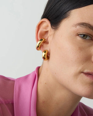 A close-up of someone wearing two gleaming 14k gold-dipped brass hoop earrings, featuring dark hair and dressed in a vibrant pink collared shirt, set against a plain, light-colored background.