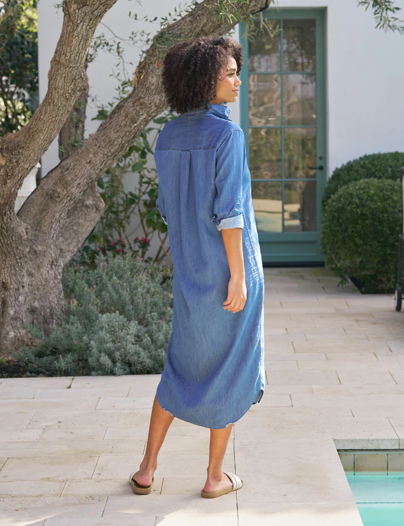A person stands outdoors on a patio, wearing a FRANK & EILEEN Rory Maxi Shirtdress in Vintage Stoneash Denim and white sandals. They have curly hair, are looking to the side with one hand in a pocket, surrounded by plants and a glass door.