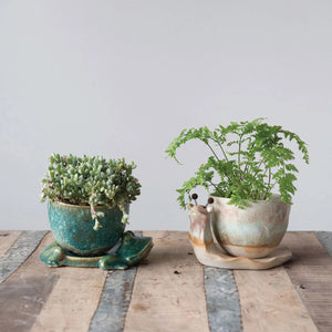 The CREATIVE COOP STONEWARE PLANTER features a green ceramic cup with textured surface, resting on a lounging frog-shaped base. The reactive glaze highlights the frog's limbs, which form part of the support, adding organic charm.