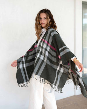 A woman with wavy brown hair drapes the MERSEA - PLAID POCKET WRAP, featuring fringe and dark stripes, over her black top and pants. This versatile travel wrap from MER SEA is ideal for any journey, providing both style and easy care as she stands against a plain light background, holding it around her shoulders.