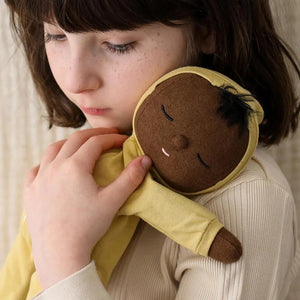 An adorable DOZY DINKUM DOLL by OLLI ELLA USA, featuring soft brown fabric skin, closed embroidered eyes, and short black hair, is dressed in a charming yellow onesie adorned with a rainbow design on the chest. Perfect as a first toy, the doll is lying on a white, quilted surface.