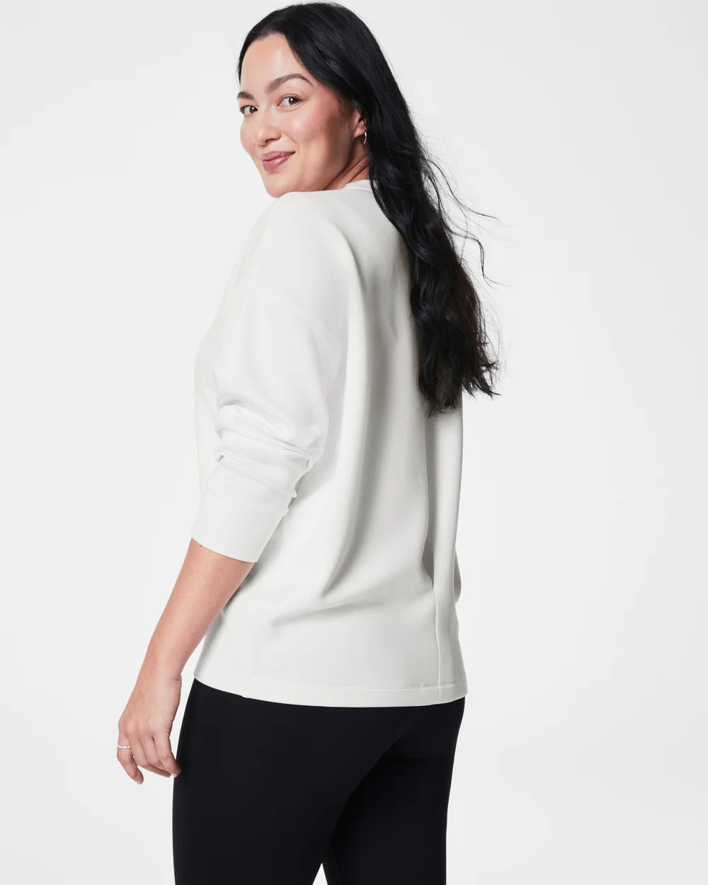 A person with long black hair stands facing slightly away from the camera, dressed in a SPANX AireEssentials Long Drew Sweater made of soft spacer fabric, complemented by black leggings and gray sneakers. The high-low hem on the sweater adds a touch of style against the plain white studio backdrop.