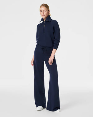 A person with short hair is wearing a SPANX AIRESSENTIALS HALF ZIP sweater in navy blue, made from lightweight spacer fabric, paired with black pants. Their arms are crossed over their chest as they stand against a plain, light grey background.