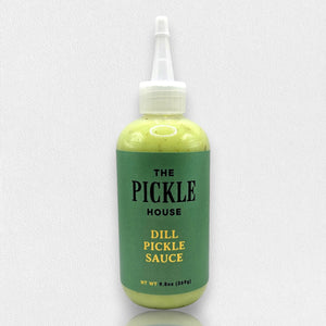 A green bottle labeled "PICKLE HOUSE - DILL PICKLE SAUCE" with a white nozzle is showcased on a plain, light gray background. The versatile 9.5 oz (269g) sauce by THE PICKLE HOUSE can be used as a salad dressing, dipping sauce, or marinade.