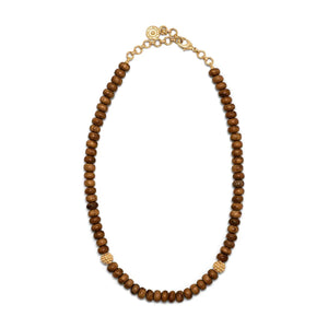 Close-up of a person wearing two necklaces. The shorter necklace, part of the CAPUCINE DE WULF - EARTH GODDESS SINGLE STRAND NECKLACE collection, has small circular wooden beads with a few textured gold beads interspersed, adding an element of earthy opulence. The longer necklace features larger hand-carved teak beads connected by 18K Gold IP over Brass loops. The person is wearing a white outfit.