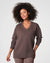 A person with curly hair is standing and looking to the side, wearing a cozy SPANX - AIRESSENTIALS BRUSHED V-NECK TUNIC paired with brown pants. The breathable top drapes elegantly, capturing a sense of laid-back comfort against the plain, light-colored background.