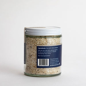 The JACOBSEN SALT - INFUSED BLACK PEPPER SALT is housed in a glass jar with a white lid. The label lists sea salt and black pepper as the ingredients and notes the absence of anti-caking agents, so natural clumping may occur. It asserts that the product is not bioengineered and touts its suitability for enhancing recipes. This product is manufactured by JACOBSEN SALT CO. in Portland, OR.