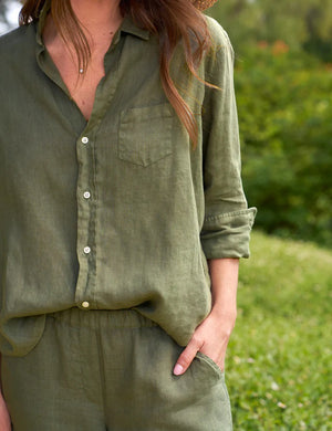 A person stands outdoors wearing an olive green FRANK AND EILEEN - EILEEN Relaxed Button Up Shirt in Linen from the Tuscany Getaway Set with the sleeves rolled to the forearms and matching Maisie pull-on pants. Their left hand is partially in their pocket, and greenery is visible in the background.